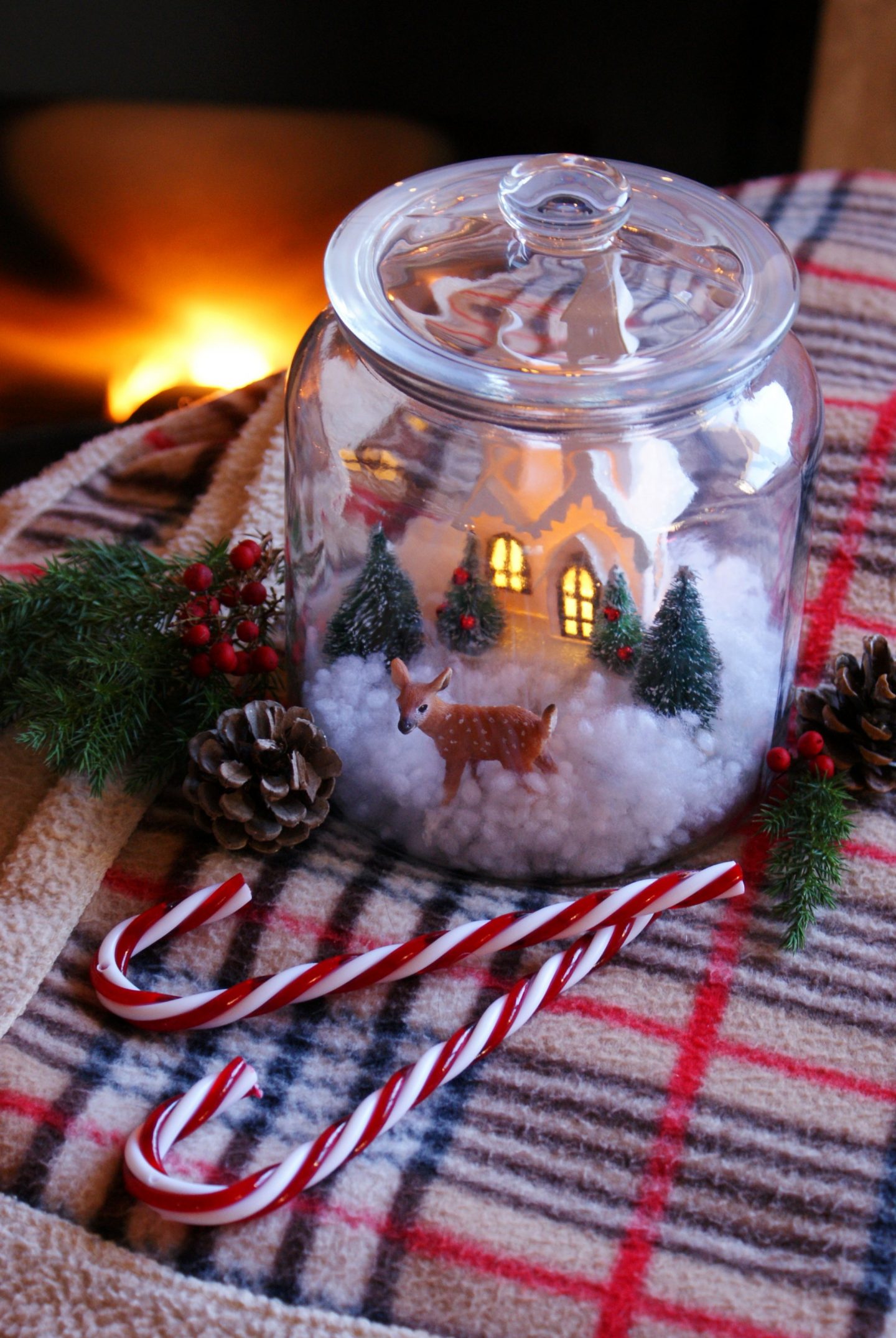 A cozy little snow scene sits inside a glass jar.  There is a small house with lights in the windows and a deer walking out front, surrounded by pine trees.  The jar sits on a fleece blanket with mistletoe, peppermints, and pinecones around it.
